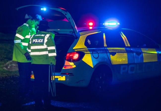 Two officers at night looking into a boot of a Police car with it's blue lights flashing