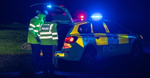 Two officers standing at rear of Police car with blue lights on at night