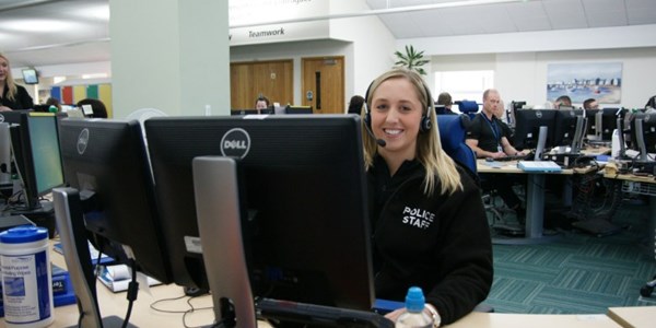 Female call handle sat at her desk in front of two screens with a head set on smiling to camera