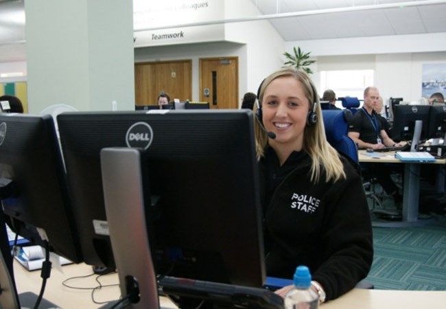 Female call handle sat at her desk in front of two screens with a head set on smiling to camera