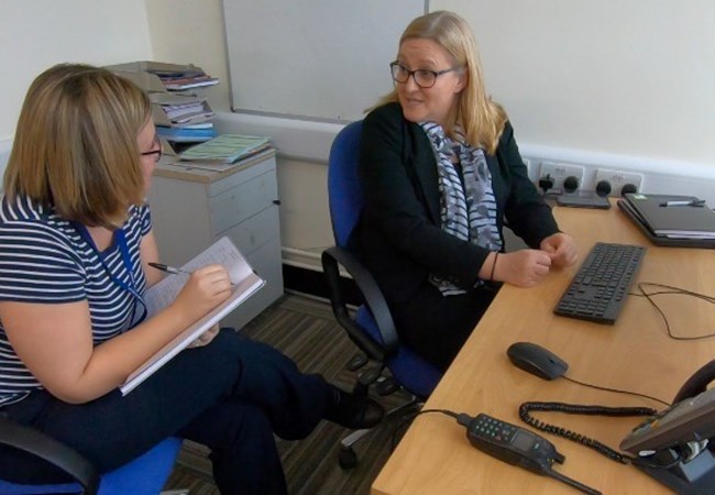 Two female detectives sat at a desk discussing a case one taking notes